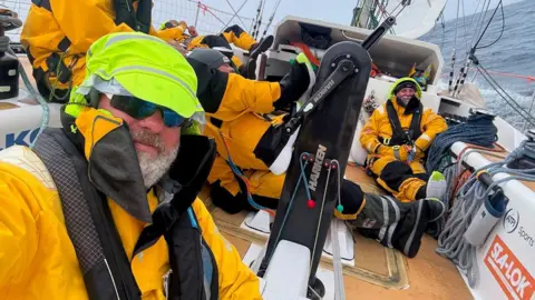 Bob is wearing waterproofs on a boat at sea. There are other crews in the background. 