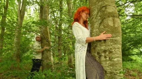 The image shows some dense woodland. A woman in the foreground is hugging the trunk of a large tree. A man in the background is hugging another large tree.
