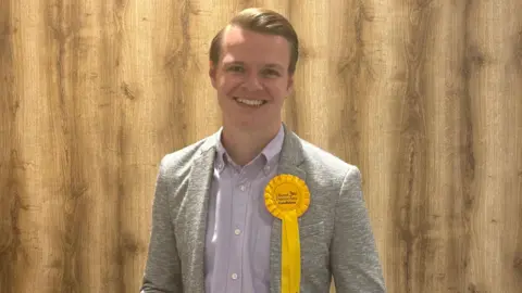 Joshua Reynolds in a pale grey suit wearing a yellow Liberal Democrats rosette smiling at the camera