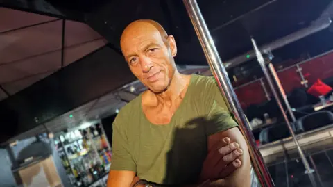 Paul Moseley/BBC Daz Crawford posing in his club next to a dancing pole. His arms are folded and he's wearing a green t-shirt.