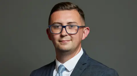 Parliamentary portrait of Crewe and Nantwich Labour MP Connor Naismith against a grey background. He has short, brushed-back brown hair, dark-rimmed glasses and is wearing a grey suit with pinstripe check, a white shirt and pale blue tie.