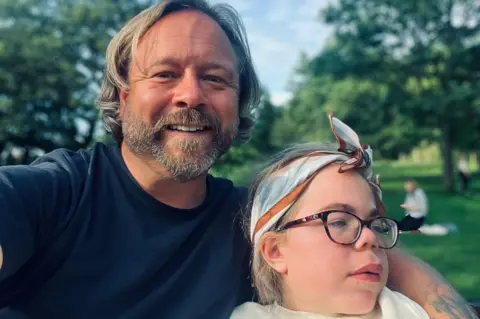 Darren Scott Darren and Sophia Scott. He has longish blond hair and a beard and is wearing a blue T‑shirt. His arm is around Sophia who is wearing glasses and a blue, brown and white cotton scarf tied around her head. He is smiling at the camera while she is looking off camera. They are in a park.