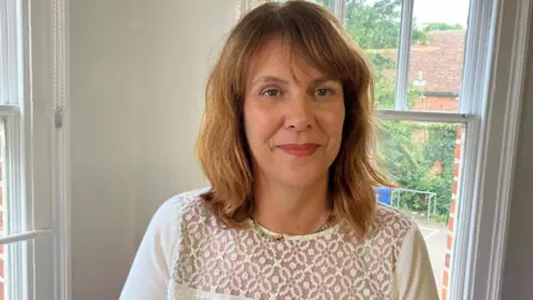 Kim Trotter is standing in a bay window. She is wearing a white jumper which has a lace detail. She is smiling. Part of a garden is visible behind her.
