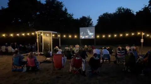 BBC An audience watches a movie projected from a horse box in a field