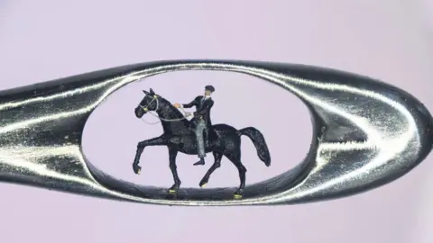 A micro sculpture of a man in a black coat, black hat and grey trousers sat in a horse inside the eye of a needle