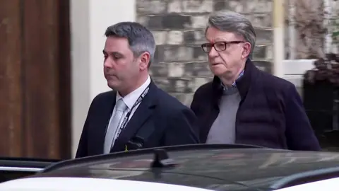 Lord Mandelson leaves his London home, walking behind a police officer wearing a suit, towards a white car with the door open.