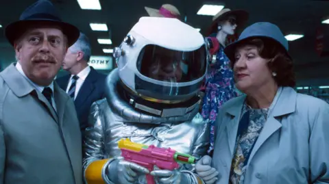 Three cast members of Keeping Up Appearances inside Northampton's former Debenhams. Richard is wearing a trench coat and hat and is looking away from the camera, embarrassed. "Daddy" is in the middle wearing a spacesuit fancy dress costume while holding a water pistol. Hyacinth is wearing a blue coat with a blue hat, and is looking at "Daddy" in an accusatory way.