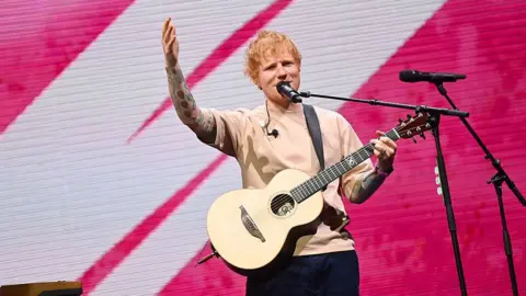 Getty Images Ed Sheeran is pictured performing on stage. He is dressed in a light-coloured t-shirt and dark jeans, holding an acoustic guitar. He is on stage with microphones in front of him and a pink and white led screen behind. 