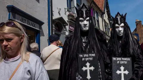 Getty Images Three people on a street of shops. On the left edge of the frame a woman is wearing a light grey sweatshirt and has sunglasses resting on the top of her head. A brown bag strap crosses her chest. To the right two costumed figures stand side by side facing the camera. Both figures are wearing dark gothic-style outfits which include long black cloaks, black crowns, white face coverings and dark eye openings. They also have long black hairpieces and black gloves.