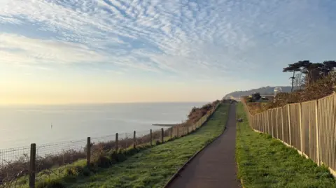 Arnie A paved footpath along a cliff top. The path is lined on each side by grass and on one side is a wooden fence. The other side is a wire fence and beyond the sea is blue. The sky has light white clouds and is lit by a low sun that is glowing yellow