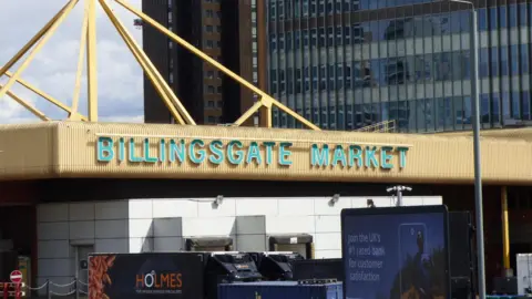 Green writing on yellow roof spells out 'Billingsgate Market' below which are lorries and behind it are tall glass buildings