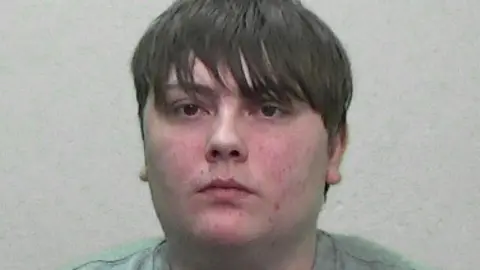 Mugshot of Elliot Robinson. He is clean shaven with a large round spotty face and greasy-looking brown hair, with a fringe below his eyebrows.