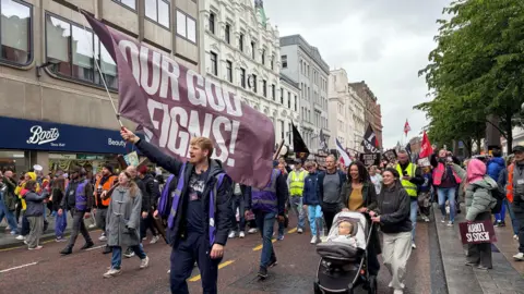 A large crowd of people march along a busy city centre street. The man at the front holds a large purple flag. He wears a purple Hi-Viz jacket, navy trousers and a navy top. 