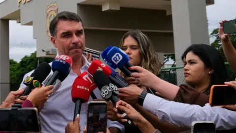 Brazil ex-President Jair Bolsonaro's surgery for hernia 'successful' REUTERS/Diego Herculano A file photo from 16 December shows Senator Flávio Bolsonaro speaking to the journalists after visiting his father at a Federal Police building in Brasilia.
He is wearing a light-blue shirt and in the background the police building can be seen. Journalists are proffering their microphones at Flávio Bolsonaro.