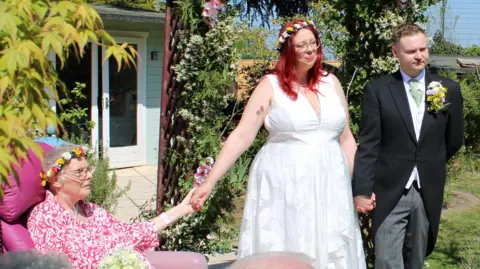 St Elizabeth Hospice A woman wearing a wedding dress holds the hand of an older woman who is sitting down on a pink chair. She has tubes come from her nose and flowers in her hair. Next to the woman is a man wearing a black suit wedding jacket, grey trousers, and a white shirt. They are looking away from the camera and are standing in a garden.