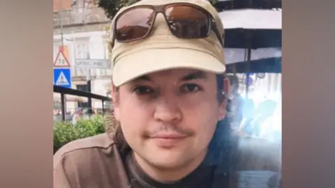Luis Piovesana shown smiling at the camera. He has dark curly hair, a moustache, and wears a light cap with darker glasses over the top.