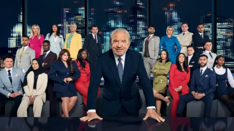 PA Media Lord Alan Sugar leans forward with both hands on a boardroom table, wearing a dark suit. Behind him, a large group of Apprentice contestants sit and stand in formal business clothing, arranged in rows with a nighttime city skyline backdrop.