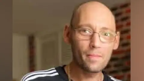 Dean Larvin is smiling slightly at the camera. He is bald with a stubbly beard. He is wearing glasses, a gold chain and a black top with white stripes on the shoulder.