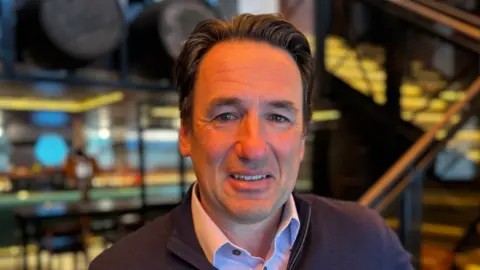 Head and shoulders shot of Ollie Vaulkhard. He is wearing a jumper over a shirt and has combed back brown hair. He is standing in one of the bars he owns in Newcastle.