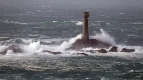 Longships Lighthouse stands tall amongst rough seas. White waves rise up against the lighthouse, and choppy blue waters can be seen in the background. It has small rocks around the base. 