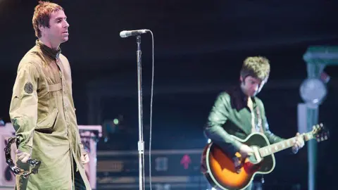 Getty Images Liam and Noel Gallagher stood on stage. Liam is wearing a green Parka coat and holding a tambourine, Noel is playing a guitar.