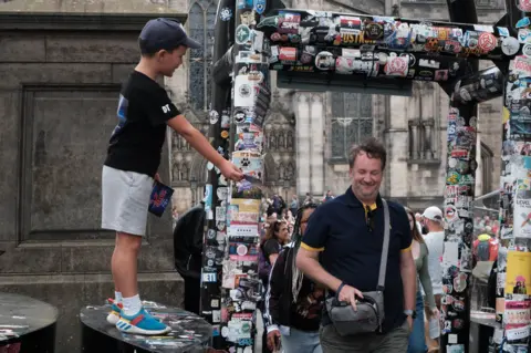 Chris Foote/BBC Scotland A young boy in shorts and a baseball cap gives out flyers standing on a street security bollard as people walk through the gate
