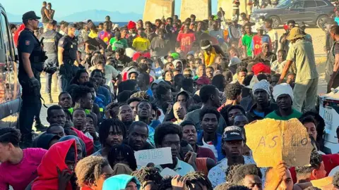 Migrants sat on a beach as police officers watch on, with some holding signs saying 'Asilo'
