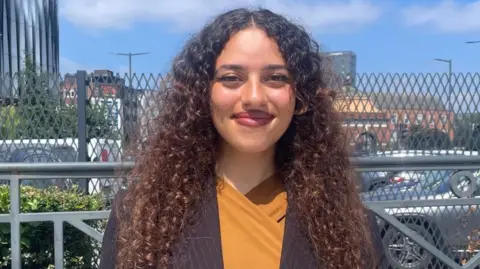 A woman with dark curly hair smiles at the camera in front of a car park wearing a mustard coloured top under a black striped blazer on a bright sunny day.