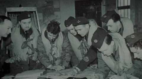 Shuan Whitmore/BBC A black and white image of eight 1940s or 1950s RAF personnel. They are bending over a chart on a table and are wearing uniforms. Most of the men have life belts on over their uniforms.