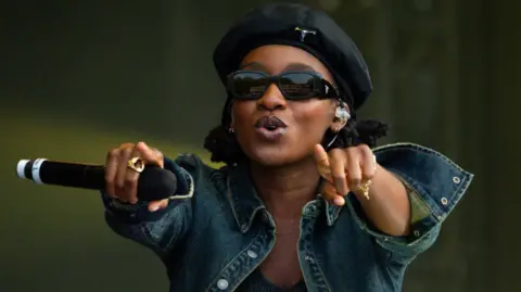 Little Simz performs on stage wearing a denim jacket, black beret and pitch black sunglasses. She is pointing to the audience and encouraging them to sing along.