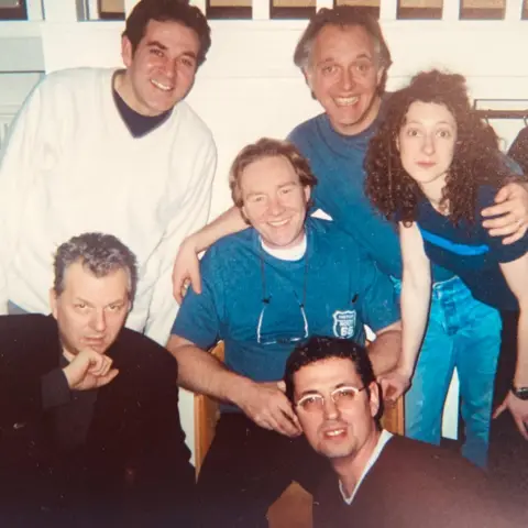 Bob Baldwin A group of six people. One man is sitting in the centre surrounded by four men (including Rik Mayall) and a woman in a behind-the-scenes photo. Mayall has his arm around the man in the centre.