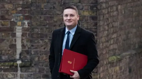 Getty Images Wes Streeting, a man with short dark hair, wearing a suit and carrying a red folder under his arm.