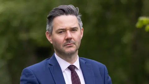 DNEAT Oliver Burwood wearing a blue suit, white shirt and purple patterned tie giving a serious look into the camera