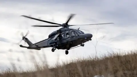 A black helicopter in flight, coming in to land on a field with long grass. 