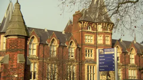 The red‑brick Mossbourne Community Academy building in Hackney, with tower features and a sign directing visitors to the main entrance.