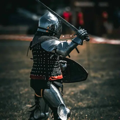 Kyle Edwards Close-up of a single person in full medieval-style metal armour standing on a grassy field. The figure holds a sword raised diagonally above shoulder height and a curved shield positioned in front of the body. The armour includes a steel helmet with a face visor, chainmail around the neck, and riveted black chest protection with small metal studs. The background is blurred, showing a roped-off arena and indistinct figures in red clothing.