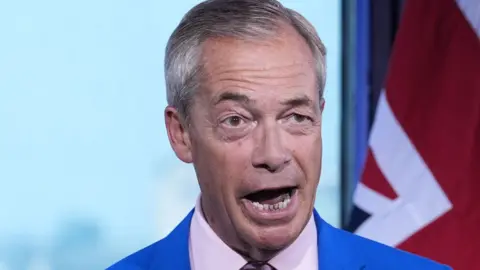 Nigel Farage, mid-way through making a statement. Only his head and shoulders are visible. He is animated, with his mouth open and one of his eyebrows is raised. Behind him on the right of the image is an out of focus Union Flag 