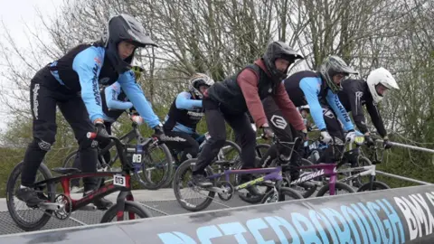 Connor Bennett/BBC At least six BMX riders poised, standing up on their pedals leaning over their bikes ready on a ramp above the track gate, ready to go. They are all wearing helmets and fleeces. Behind them are leafless trees. 