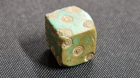 Discover Bucks Museum A close up of a bone die. The cube shaped object has gold dots on its side. 