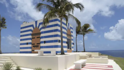 US Department of Justice A blue-and-white striped building on Little St James, one of two Caribbean islands owned by Epstein, surrounded by palm trees, set on a raised area with flat benches around. In the background is the sea and a blue sky with a few clouds.
