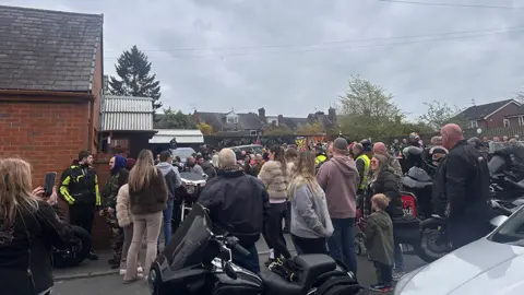 Hundreds of people outside a crematorium, with motorbikes in hte foreground