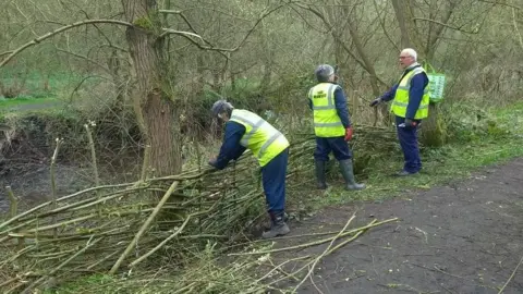 Friends of Holywell Dene Three workers in yellow hi-vis jackets are making a fence out of willow sticks in a woodland.
