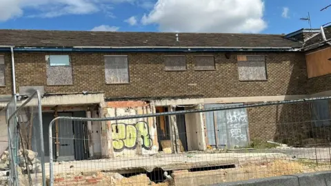 A two-storey building which has been badly vandalised with metal fencing outside. Graffiti has been sprayed on boards put in place of the windows.