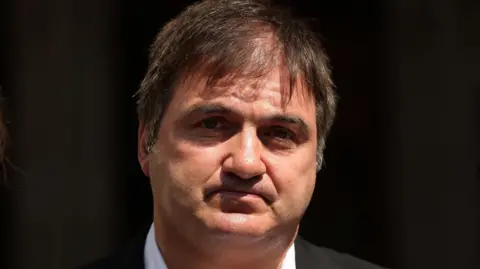 Barry George, dressed in a suit looks at the camera. He has black hair