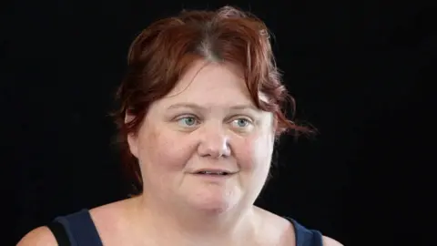 Shaun Whitmore/BBC A woman with short auburn hair looks slightly off camera, mid-speech. She has blue eyes. The image is of her head and shoulders, with a black background. The straps of a dark blue vest top are on her shoulders. 