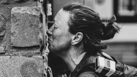 Montane Summer Spine A black and white image of a woman with her hair tied back kissing a stone wall