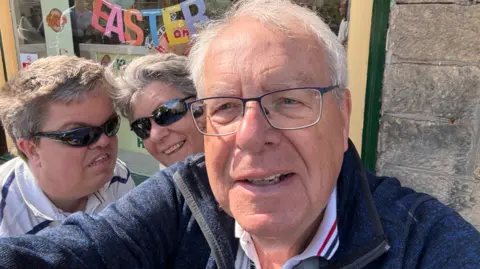 A selfie of two men and a woman in front of a shop window with a sign reading happy easter