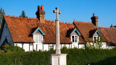 Getty Images A small war memorial - with a cross on the top - in Dorchester on Thames in Oxfordshire. the memorial is in front of a lush green hedge and a row of Tudor homes behind that are white with black wooden beams and brown tiled roofs. the sky is bright blue 