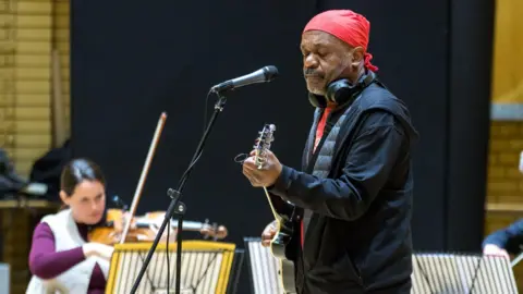 A man with a red head covering and headphones stands holding a guitar in front of a microphone. A woman sits in the back playing a violin.