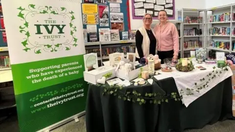 Two women manning a charity stand in a library with a banner reading The Ivy Trust supporting women who have experienced the death of a baby